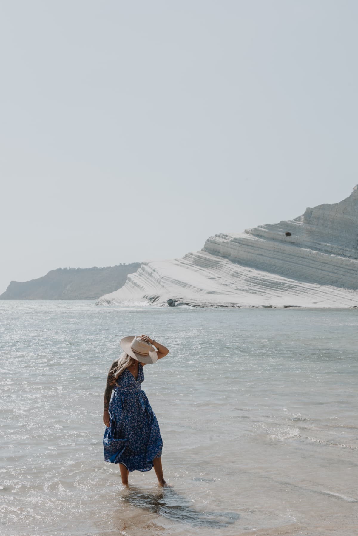 A woman in a blue dress standing in shallow water below white cliffs, edited with the Alba preset