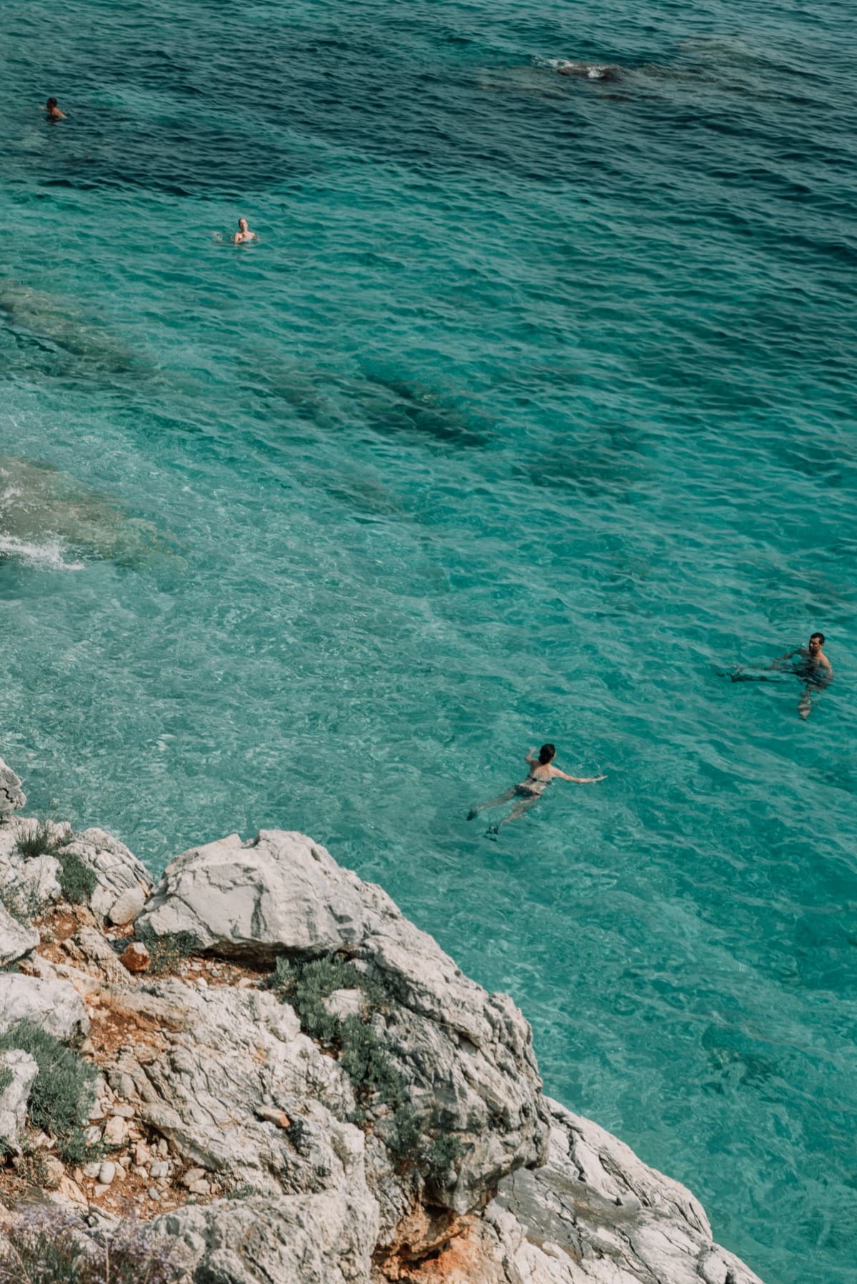 Swimmers in clear turquoise water below a rocky headland, edited with the Azzurro preset