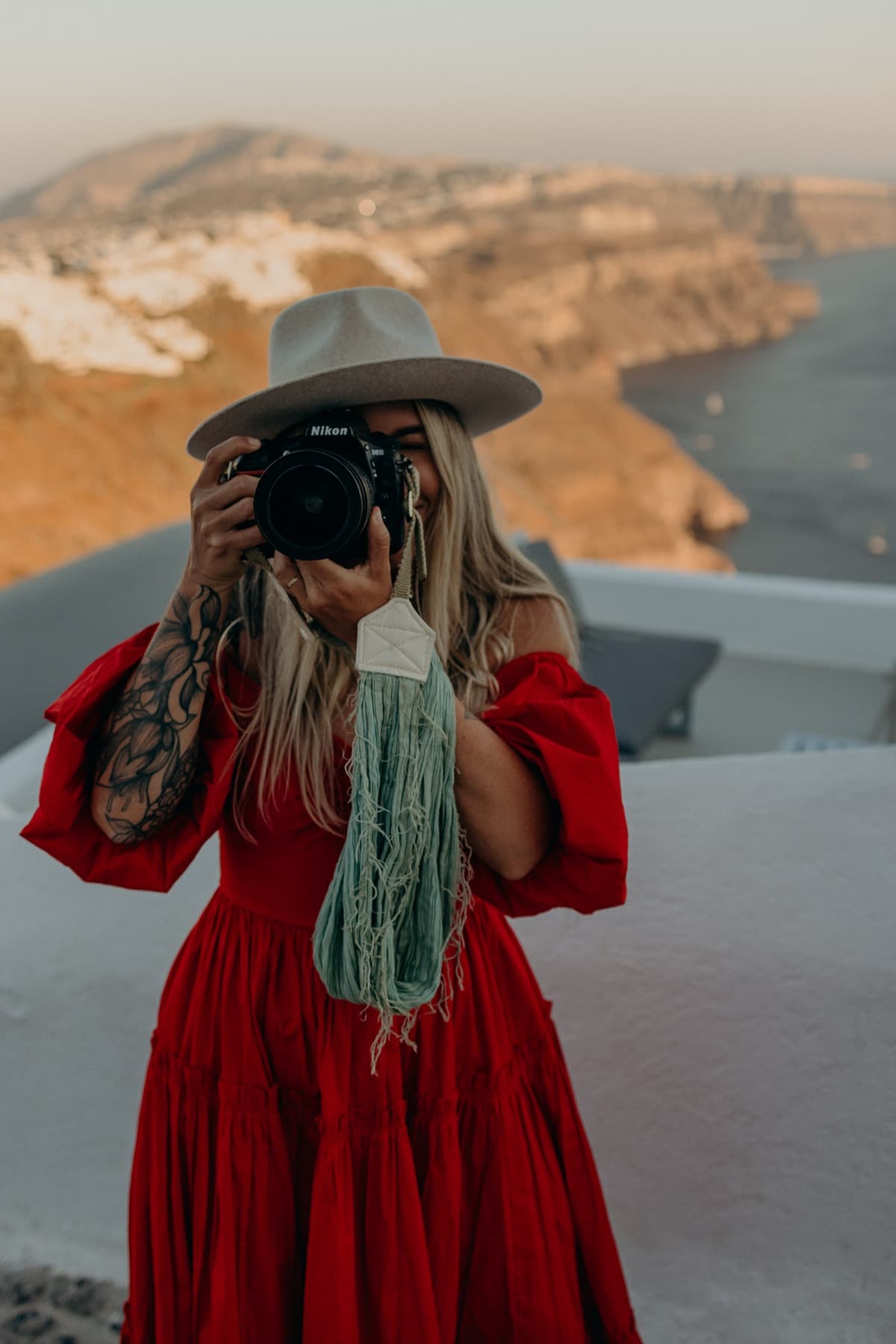 Kat Moore in a red dress, camera raised, on a terrace above the sea