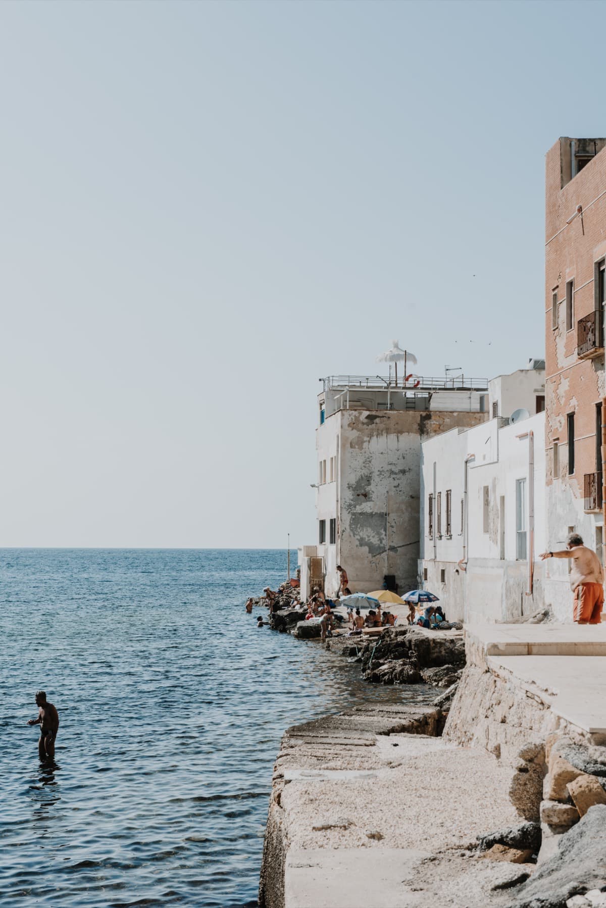 A swimmer slipping into calm water below the salmon facades of a seafront town, edited with the Mandorla preset