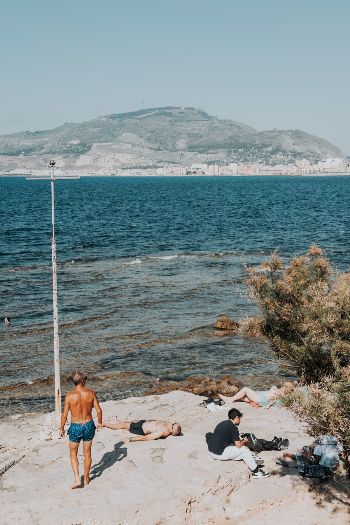 A man walking down to the water past sunbathers on a rock ledge, edited with the Sole preset